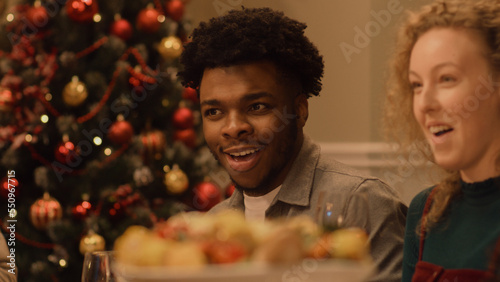 Happy multi cultural family celebrating Christmas or New Year 2023. African American man eats and smiles. Served holiday table with dishes. Warm atmosphere of family Christmas dinner at home.