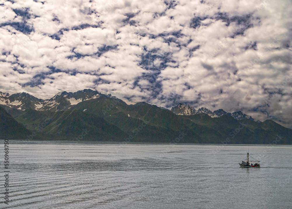 Seward, Alaska, USA - July 22, 2011: Wide landscape with ocean water and green mountain range under blue cloudscape. Small red-green fishing vessel adds colors