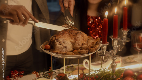 Close-up shot of African American man cutting turkey or chicken. Multi cultural family celebrating Christmas or Thanksgiving Day. Served table with dishes and candles. Family Christmas dinner at home.