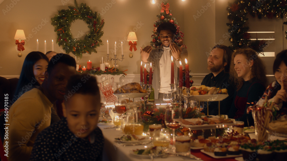 African American man givs gift to young boy. Happy family celebrating Christmas or New Year 2023. Served holiday table with dishes and candles. Warm atmosphere of family Christmas dinner at home.