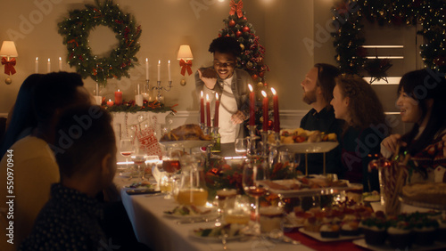 African American man givs gift to young boy. Happy family celebrating Christmas or New Year 2023. Served holiday table with dishes and candles. Warm atmosphere of family Christmas dinner at home.