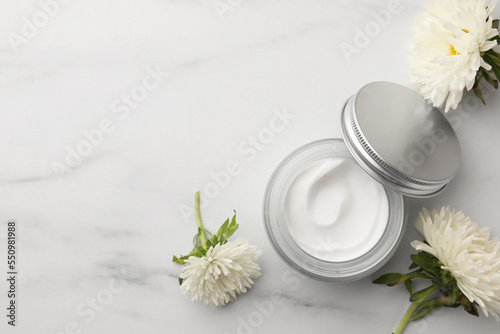 Glass jar of face cream and flowers on white marble table, flat lay. Space for text