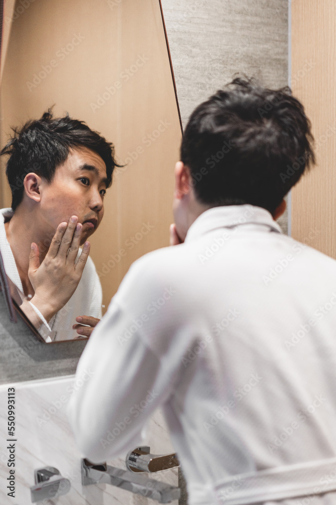 Asian man wearing bathrobe is looking himself in the mirror. Asian man wearing bathrobe is looking himself in the mirror.