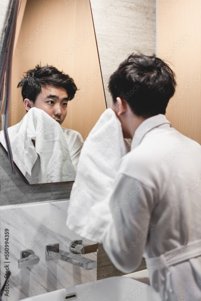 Fototapeta premium Asian man wearing bathrobe is washing face with towel in front of the mirror.