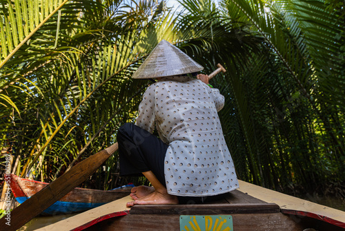 Ho Chi Minh City, Vietnam- November 9, 2022:Woman with conical Vietnamese hat controls a canoe. Tourism rowing boat in the Mekong delta. Tours on Paddle boats, tourist attraction on the Mekong River
