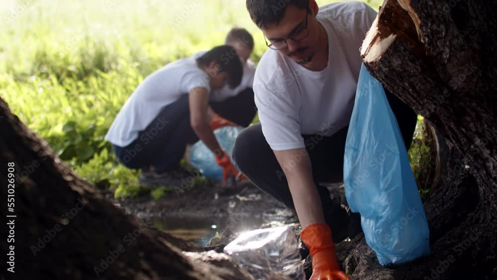 Close-up of a young guy with glasses collecting garbage in a plastic bag in nature, smiling and looking at the camera.The concept of green life and ecology Close-up of a young guy with glasses collecting garbage in a plastic bag in nature, smiling and looking at the camera.The concept of green life and ecology