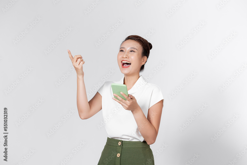 Overjoyed smiling happy cute Asian young lady point finger up hold phone posing isolated on white background. Overjoyed smiling happy cute Asian young lady point finger up hold phone posing isolated on white background.