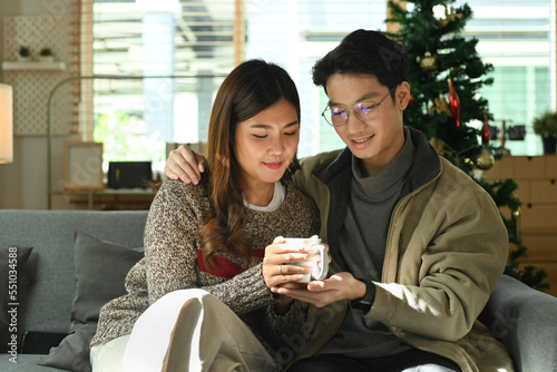 Romantic asian couple relaxing on comfortable couch, drinking hot chocolate and enjoying Christmas morning at home