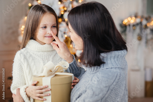 Affectionate mother gives present to her adorble little daughter, prepares surprise on Christmas, touch her nose, expresses great love. Family, celebration, presents, miracle, winter holidays concept