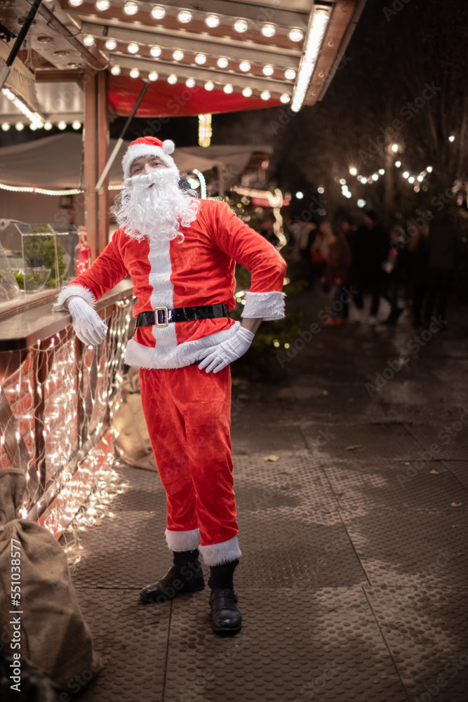 Portrait von einem verkleideten Weihnachtsmann, der auf einem Weihnachtsmarkt von Lichtern umgeben ist  zur blauen Stunde 
