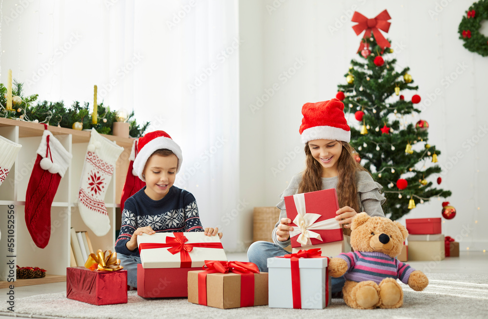 Excited boy and girl siblings in Santa hats have fun open Christmas presents together near fir-tree. Happy children enjoy unpacking New Year gifts in morning at home. Winter holiday celebration.