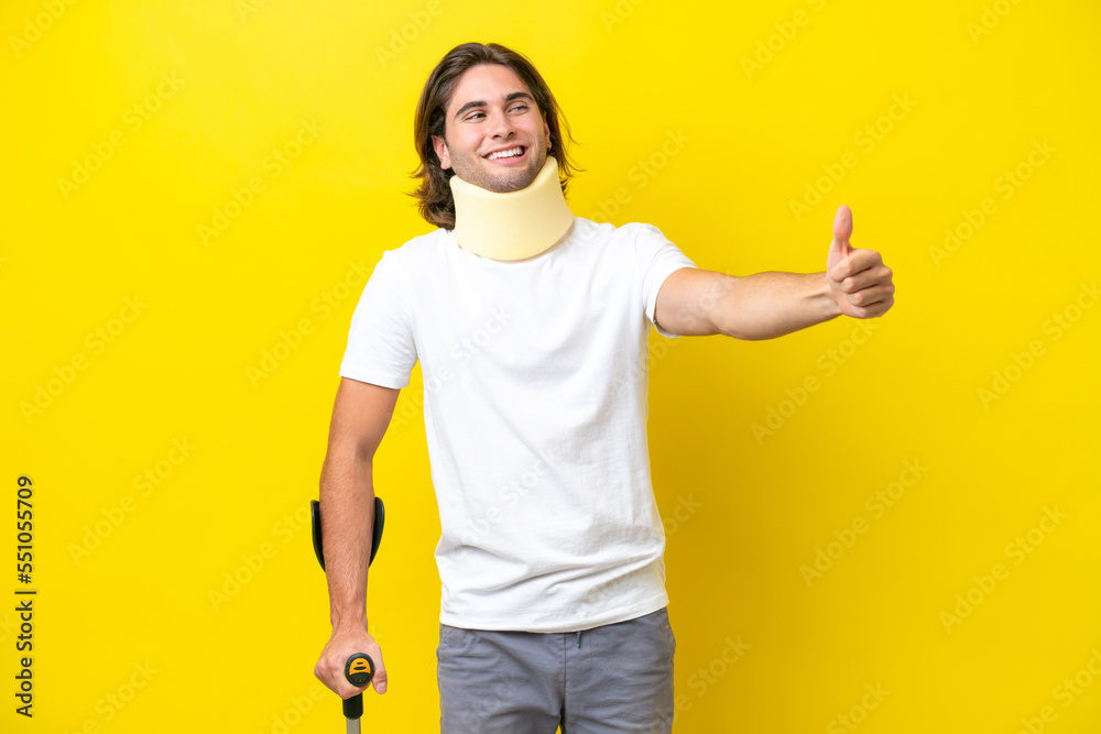 Young handsome man wearing neck brace and crutches isolated on yellow background giving a thumbs up gesture Young handsome man wearing neck brace and crutches isolated on yellow background giving a thumbs up gesture