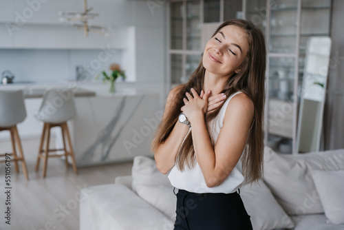 Fotografija Attractive Swedish young adult woman in white blouse and black pants folded hands on chest eyes closed feel gratitude enjoys weekend at new home