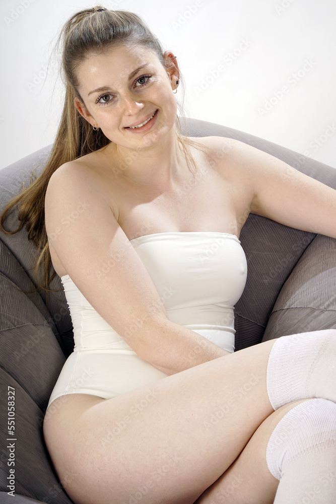 Beautiful young woman in white off shoulder body suit and stockings sits on a sofa in studio Beautiful young woman in white off shoulder body suit and stockings sits on a sofa in studio