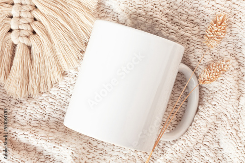 Mockup of white mug on knitted plaid with macrame decor and dried flowers, neutral color. Blank coffee cup mug mock up in Boho style, flat lay.