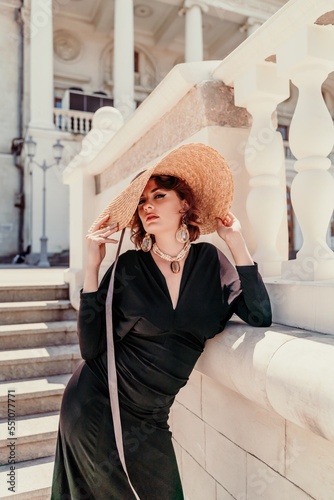 Fashion photo of a beautiful model in an elegant hat and black dress posing against the backdrop of a building with columns.