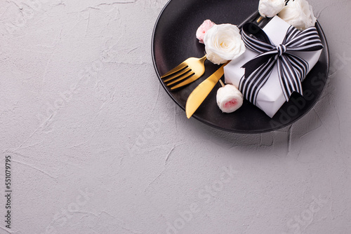 Festive table setting. Black plate, knife, fork, box with present and rwhite rose flowers on textured grey background. Top view. Place fr text. Romantic postcard. photo