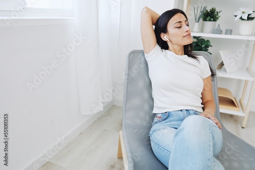Woman sitting in a chair listening to music on wireless headphones at home in jeans and a white T-shirt, fall lifestyle comfort