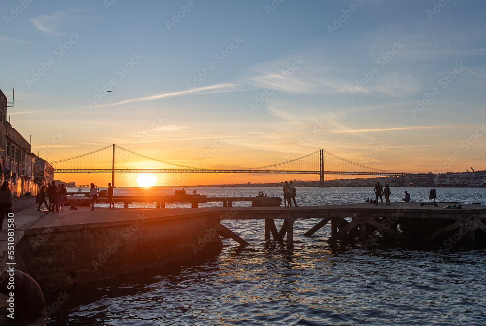 People watching the sunset in Cacilhas and Lisbon's April 25 Bridge, dock on the Tagus River and plane in the sky.