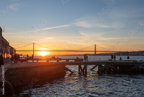 People watching the sunset in Cacilhas and Lisbon's April 25 Bridge, dock on the Tagus River and plane in the sky.