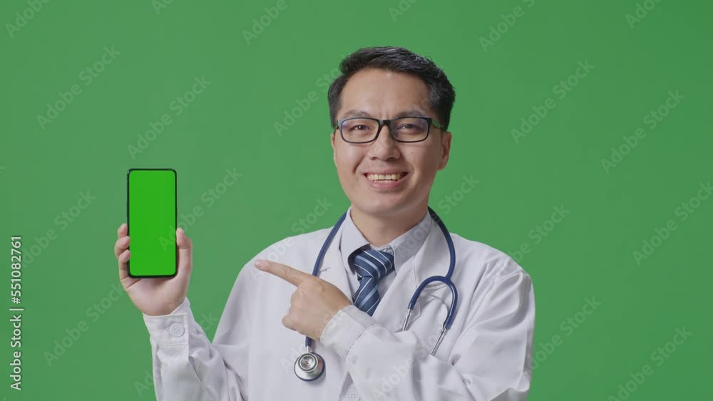 Close Up Of Asian Male Doctor With Stethoscope Pointing To Green Screen Smartphone In Hand While Standing On Green Screen Background In The Hospital
