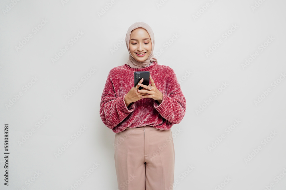 Fototapeta premium A portrait of a happy Asian woman wearing a pink sweater and hijab, holding her phone, isolated by white background