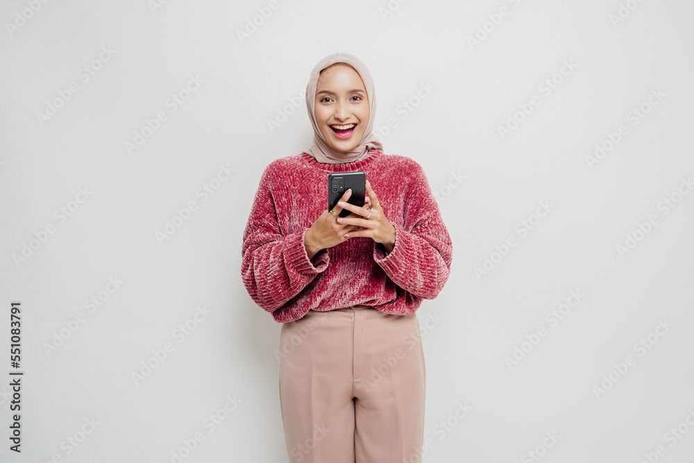 Fototapeta premium A portrait of a happy Asian woman wearing a pink sweater and hijab, holding her phone, isolated by white background