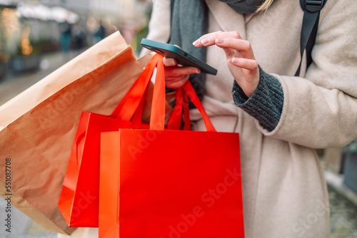 Cropped image of female hands holding red holiday gifts packages used phone on the city street near the shopping center. Retail sale concept. Shopper. Sales. Shopping Center.Cyber Monday.Black Friday