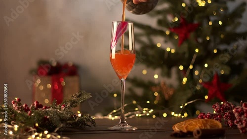 The bartender makes Christmas cocktails. An orange cocktail is poured into a glass. Christmas tree with garlands and a gift in the background. Sliced orange, cinnamon, star anise, red berries on the