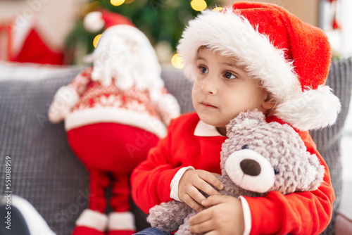 Adorable hispanic toddler hugging teddy bear sitting on sofa by christmas tree at home