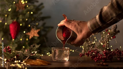 The bartender makes Christmas cocktails. A pink drink is poured into a glass through a sieve from a shaker. Christmas tree with garlands and a gift in the background. Sliced orange, cinnamon, star