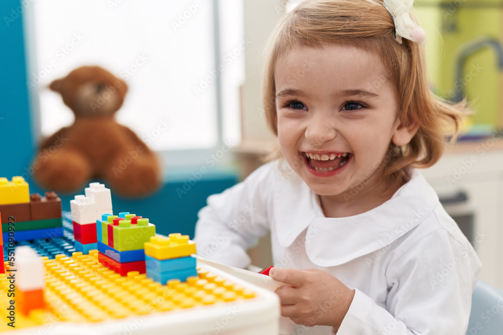 Fototapeta premium Adorable caucasian girl playing with construction blocks sitting on table at kindergarten