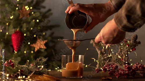 The bartender makes Christmas cocktails. Coffee cocktail is poured from a shaker into a glass with ice. Christmas tree with garlands and a gift in the background. Sliced orange, cinnamon, star anise