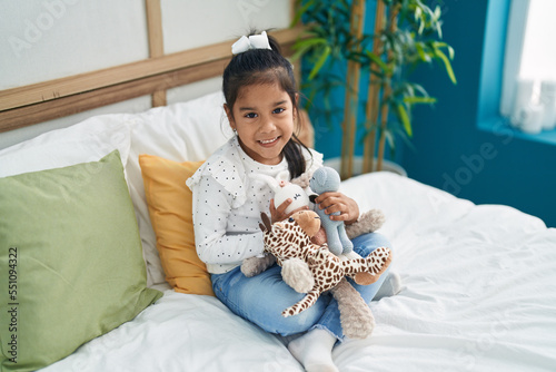 Adorable hispanic girl holding dolls sitting on bed at bedroom