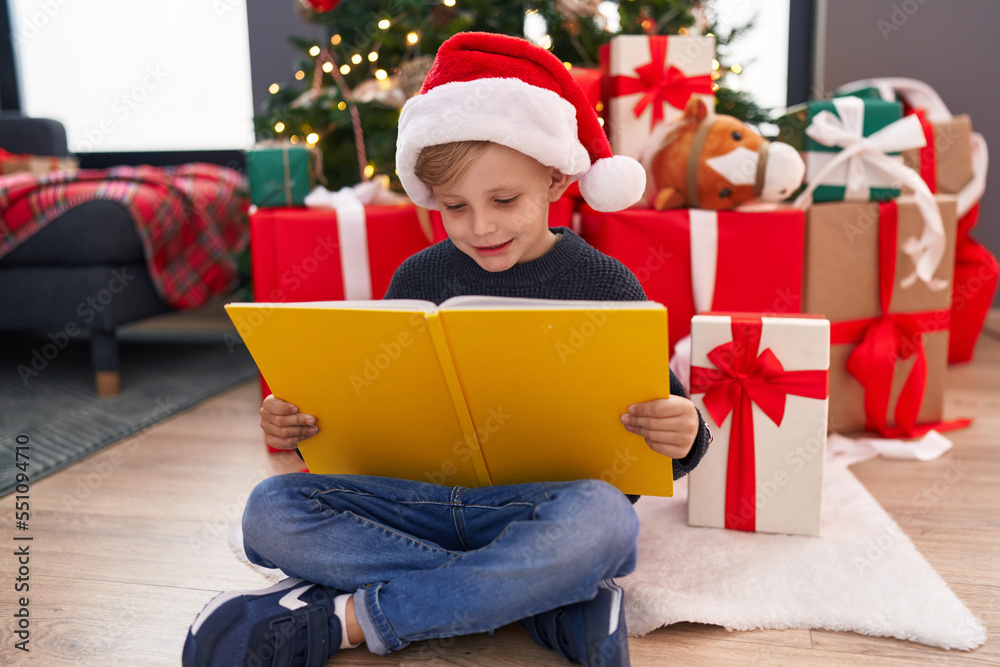 Adorable toddler reading book sitting by christmas tree at home Adorable toddler reading book sitting by christmas tree at home