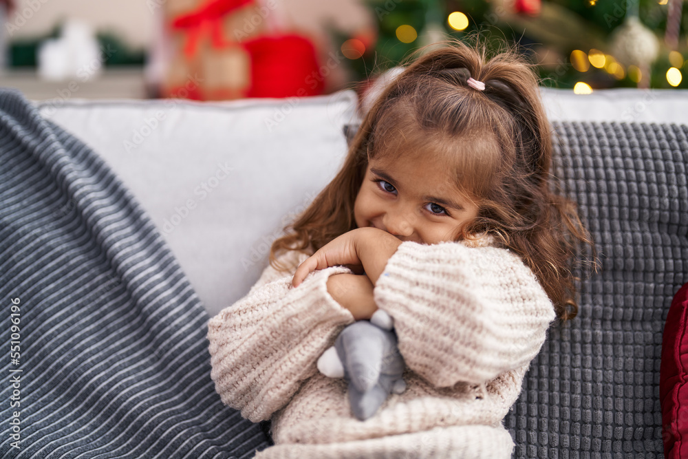 Adorable hispanic girl holding doll sitting on sofa by christmas tree at home Adorable hispanic girl holding doll sitting on sofa by christmas tree at home