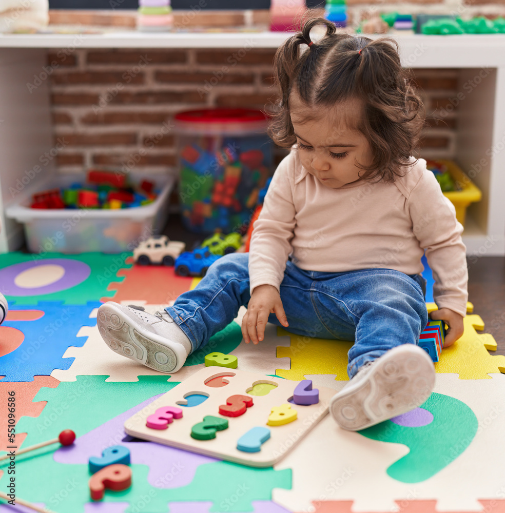 Adorable hispanic girl playing with maths puzzle game sitting on floor at kindergarten Adorable hispanic girl playing with maths puzzle game sitting on floor at kindergarten