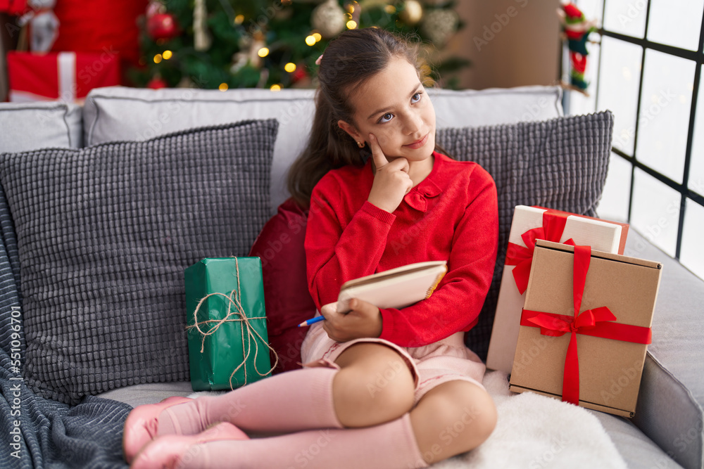 Adorable hispanic girl holding notebook sitting on sofa by christmas tree with doubt expression at home Adorable hispanic girl holding notebook sitting on sofa by christmas tree with doubt expression at home