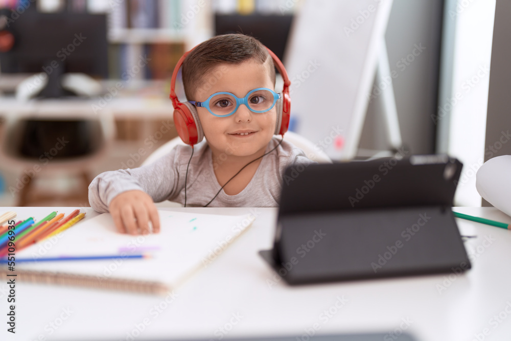 Adorable hispanic toddler student using touchpad and headphones studying at classroom Adorable hispanic toddler student using touchpad and headphones studying at classroom