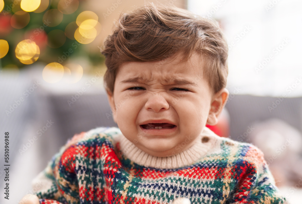 Adorable hispanic toddler sitting on sofa by christmas tree crying at home Adorable hispanic toddler sitting on sofa by christmas tree crying at home