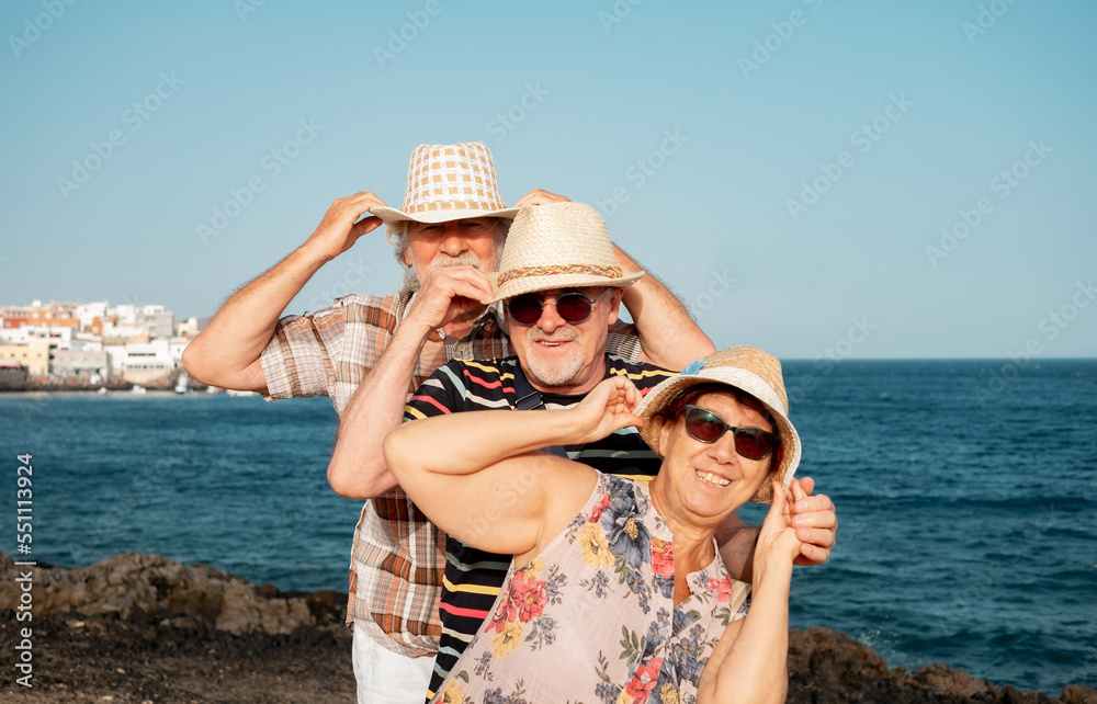 A group of three playful older people holding their hats from flying off enjoying a windy day at sea. Happy retirees on beach vacation. Horizon over water A group of three playful older people holding their hats from flying off enjoying a windy day at sea. Happy retirees on beach vacation. Horizon over water
