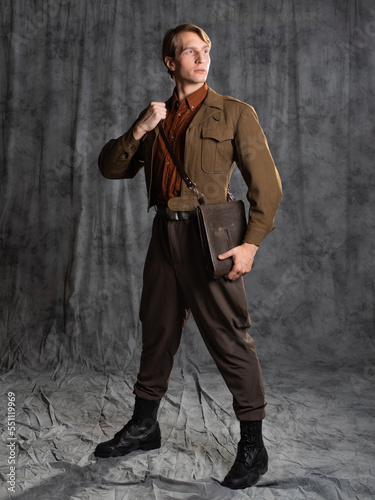 A young guy in military-style clothes, a brown flight jacket and breeches with suspenders. Posing in the studio on a gray background