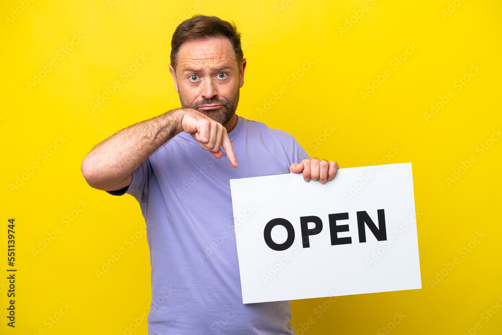 Middle age caucasian man isolated on yellow background holding a placard with text OPEN and pointing it Middle age caucasian man isolated on yellow background holding a placard with text OPEN and pointing it