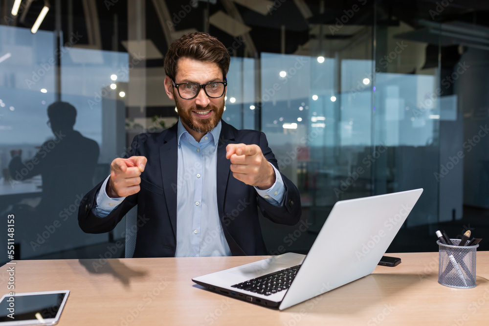 Successful mature businessman looking at camera and pointing finger forward calling to action, investor in business suit inside modern office working on laptop, boss with beard and glasses. Successful mature businessman looking at camera and pointing finger forward calling to action, investor in business suit inside modern office working on laptop, boss with beard and glasses.