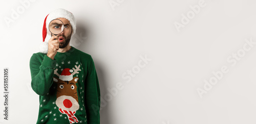 Christmas and holidays concept. Curious guy looking at something through magnifying glass, standing in Santa hat with xmas sweater, standing over white background