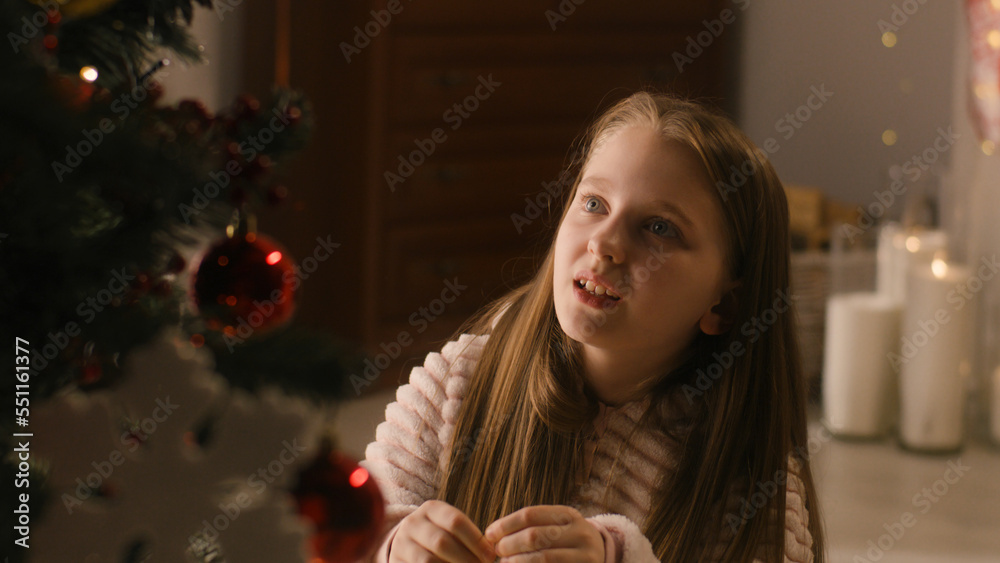 Young girl in mild pajama sits on the floor, decorates Christmas tree with balls and toys, smiles and spends leisure time at home. Child prepares room before winter holidays and New Year. Slow motion. Young girl in mild pajama sits on the floor, decorates Christmas tree with balls and toys, smiles and spends leisure time at home. Child prepares room before winter holidays and New Year. Slow motion.