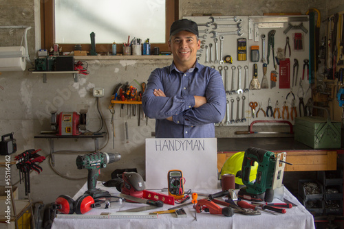 Image of a smiling man in his workshop with a bench full of work tools and the word handyman. Man capable of repairing and fixing everything in the house 