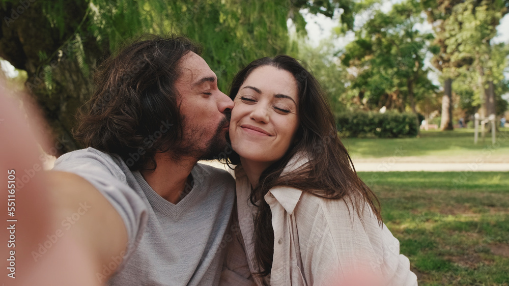 Happy couple kissing and taking a selfie sitting on blanket in park Happy couple kissing and taking a selfie sitting on blanket in park