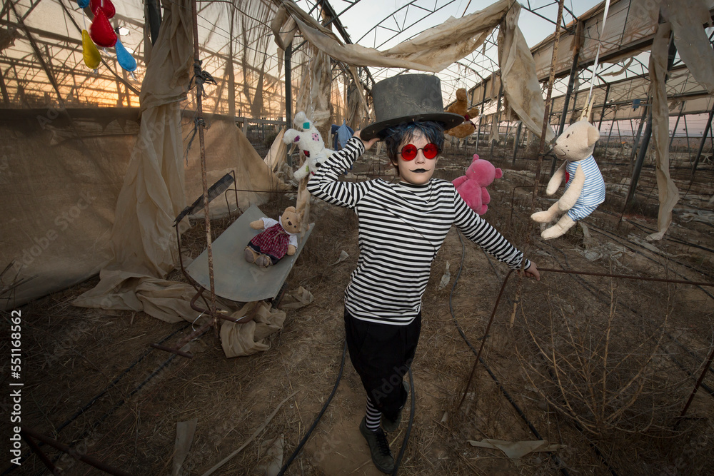 Fototapeta premium a boy in red sunglasses, a scary smile in a clown costume and make-up plays with an old cart, in an abandoned greenhouse among hanging toys