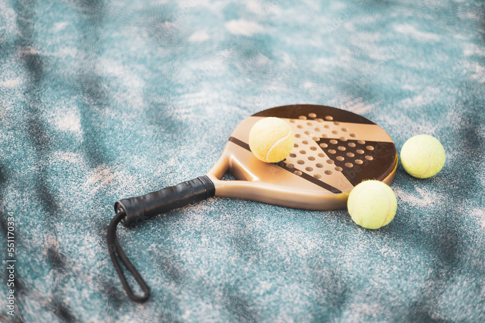 A paddle racket resting on the ground with 3 balls, the racket is seen behind the net.